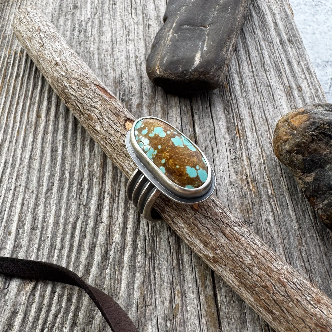  A turquoise and silver ring with a four-line band, displayed on a wooden surface with rocks and a leather lace in the background.