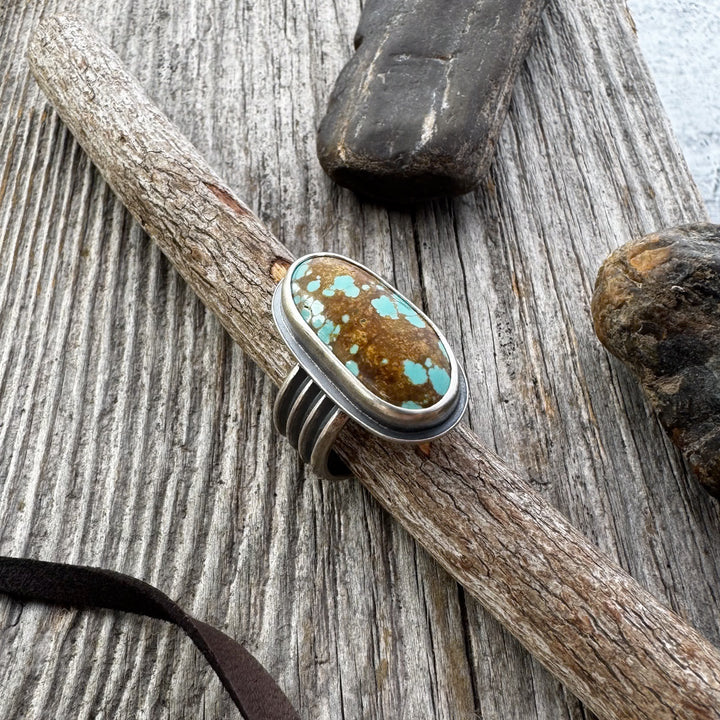  A turquoise and silver ring with a four-line band, displayed on a wooden surface with rocks and a leather lace in the background.