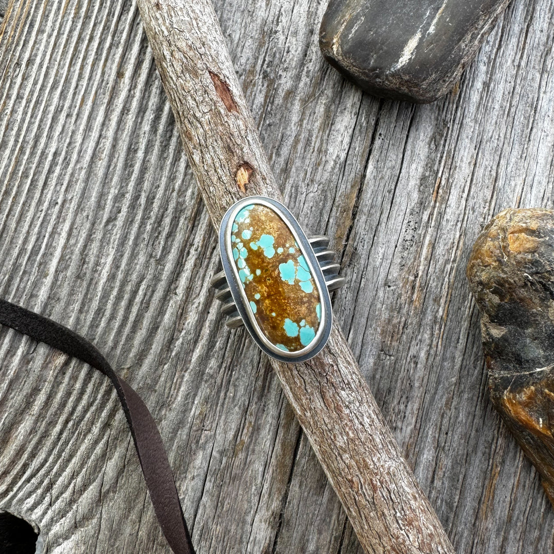A turquoise and silver ring with a four-line band, displayed on a wooden surface with rocks and a leather lace in the background.
