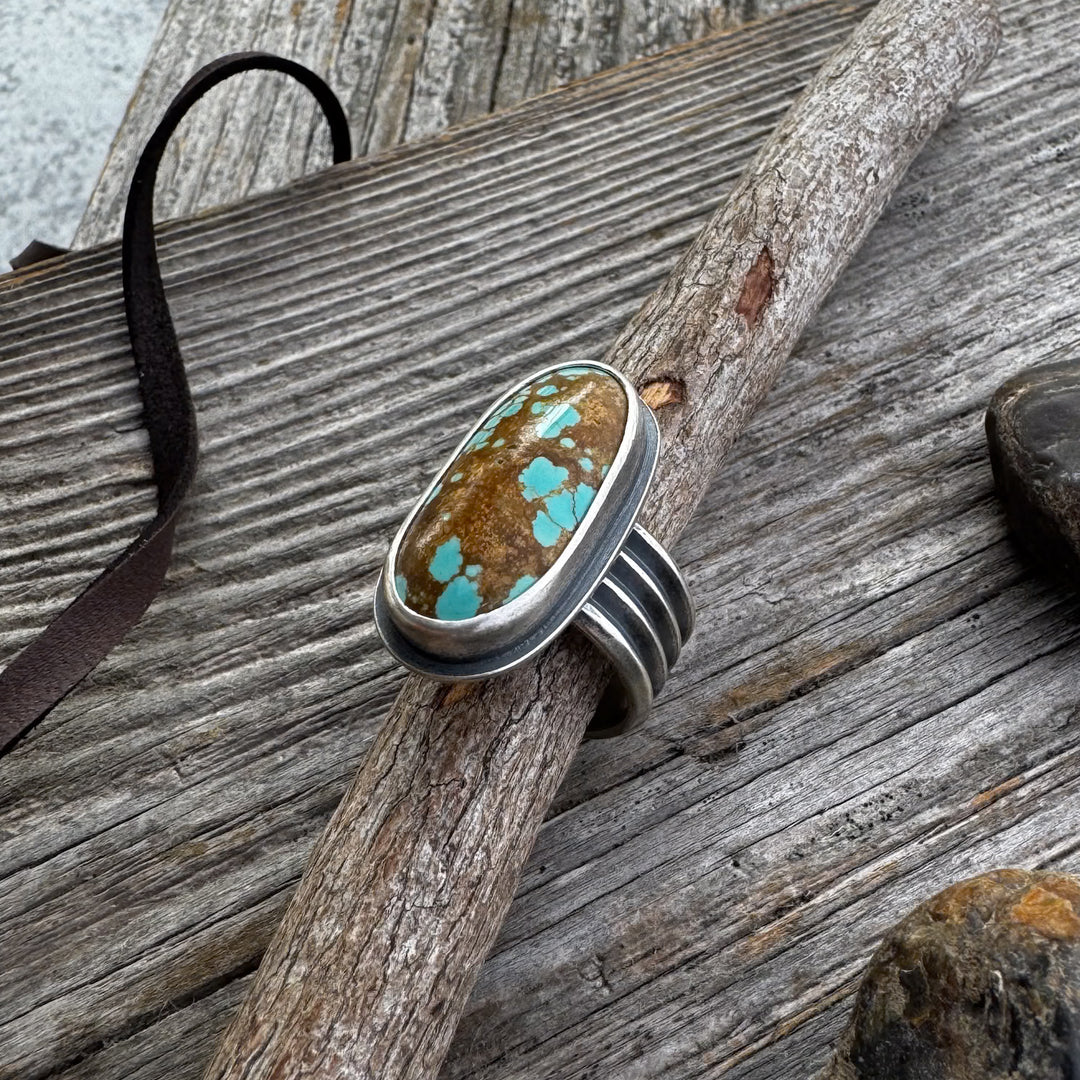 A turquoise and silver ring with a four-line band, displayed on a wooden surface with rocks and a leather lace in the background.