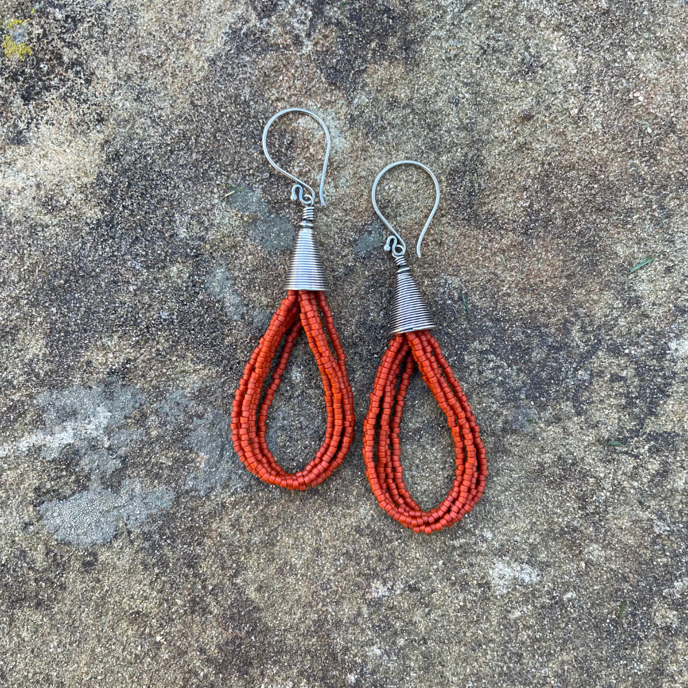 Orange seed bead earrings with sterling silver ear wires and Bali-style cone tops displayed on a textured stone background.