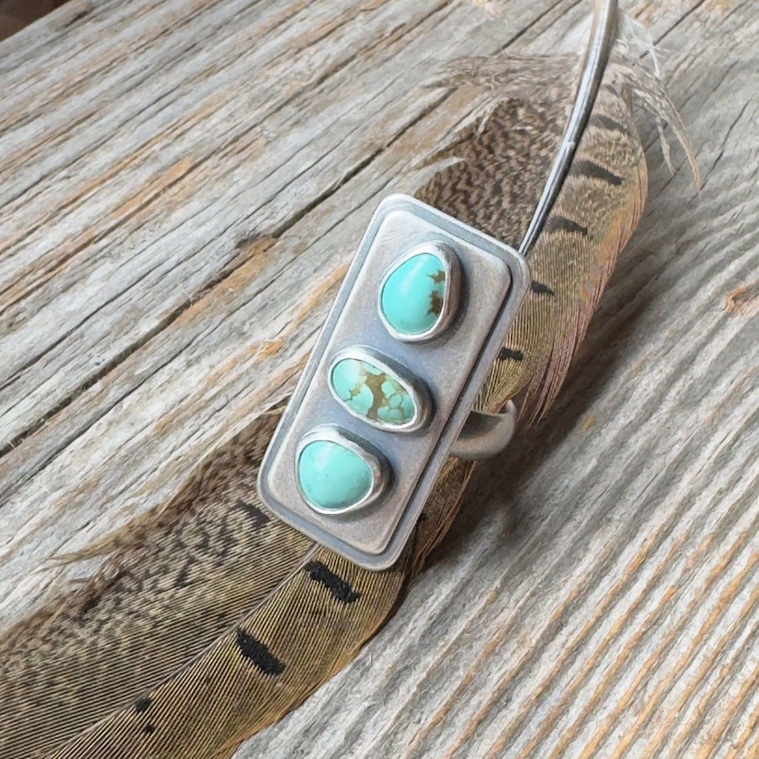 Three stone #8 turquoise sterling silver statement ring with brushed oxidized finish, size 9 on wood background with feather.