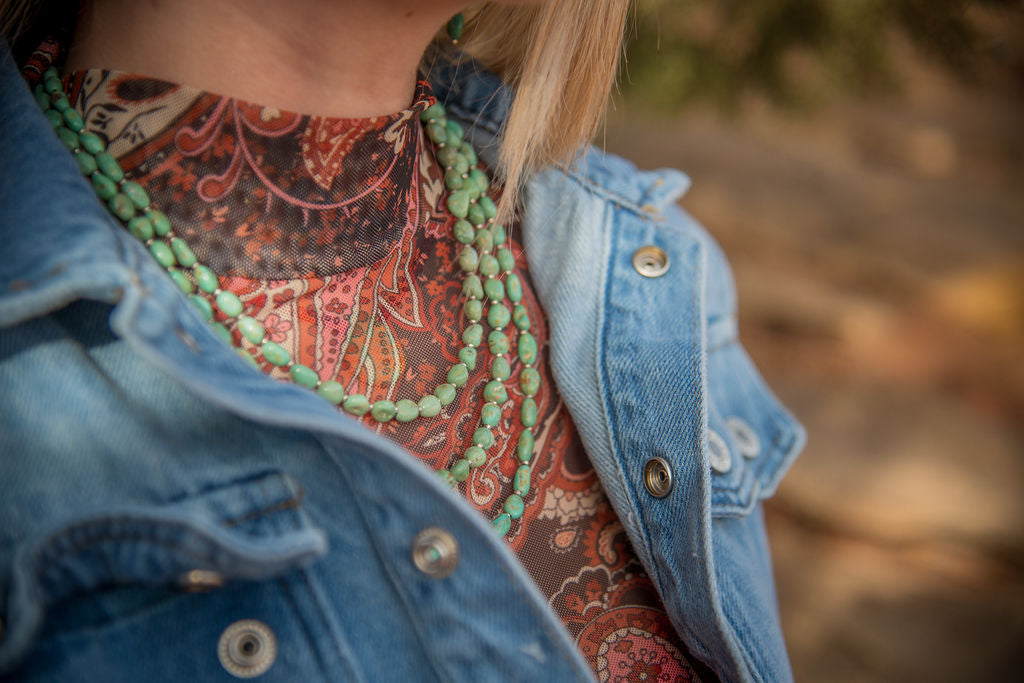 Person wearing a denim jacket over a patterned shirt with an Elisa turquoise nugget necklace trio, blurred background
