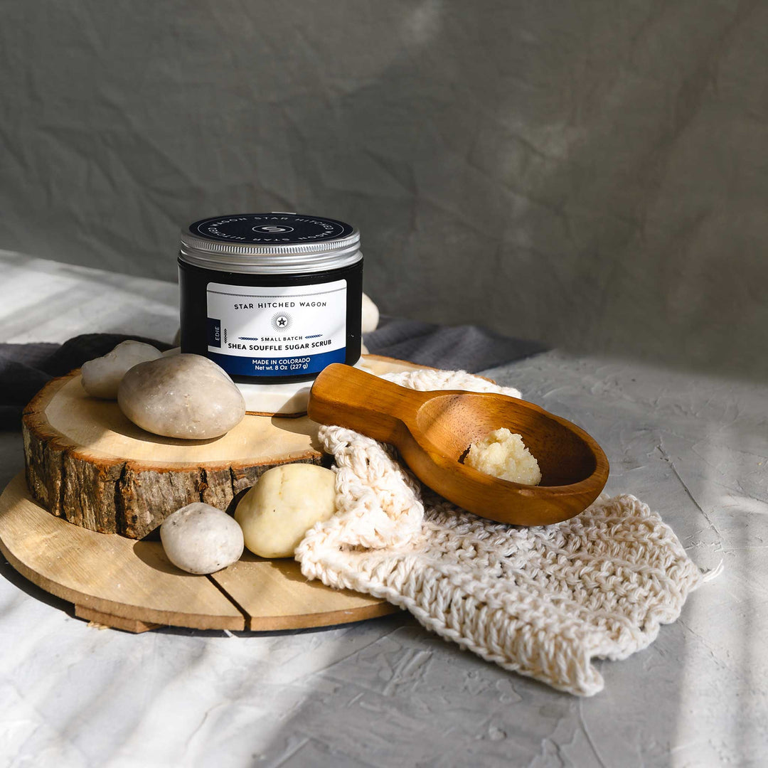 An 8 ounce jar of Edie Shea Souffle Sugar Scrub is displayed on a wooden surface, accompanied by a pebbles, wooden scoop and a knit wash cloth.