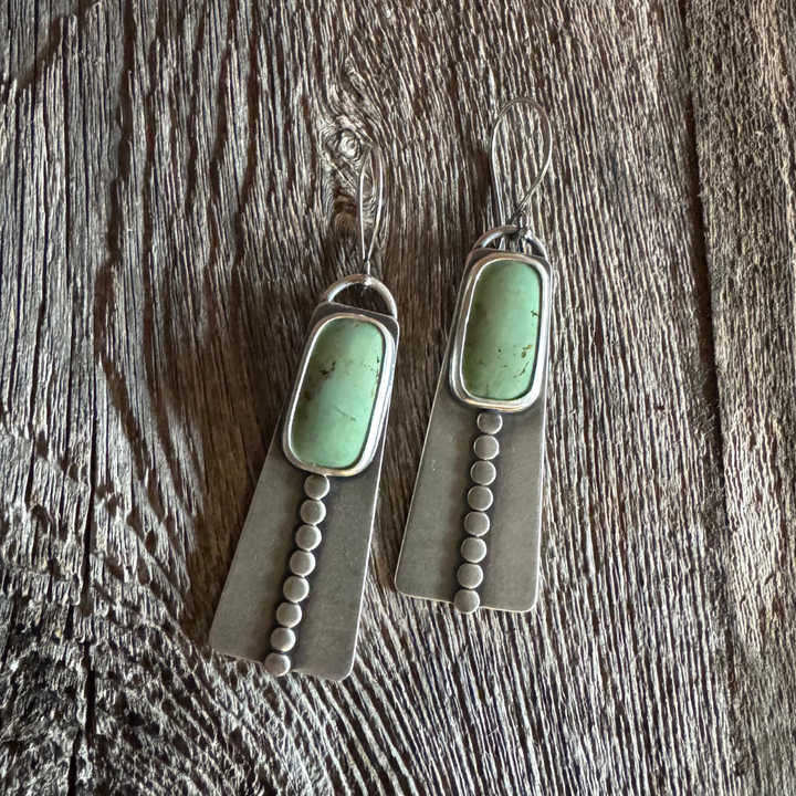 Close-up of handmade long turquoise earrings featuring green Elisa turquoise stones, sterling silver disc accents, and French hook earwires. Rustic wood and feather background .