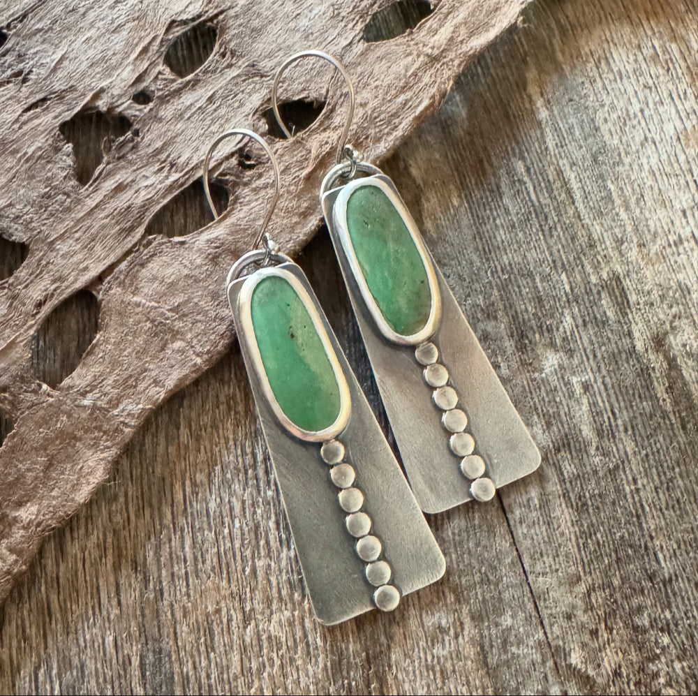 Close-up of handmade long turquoise earrings featuring green Elisa turquoise stones, sterling silver disc accents, and French hook earwires. Rustic wood and feather background .