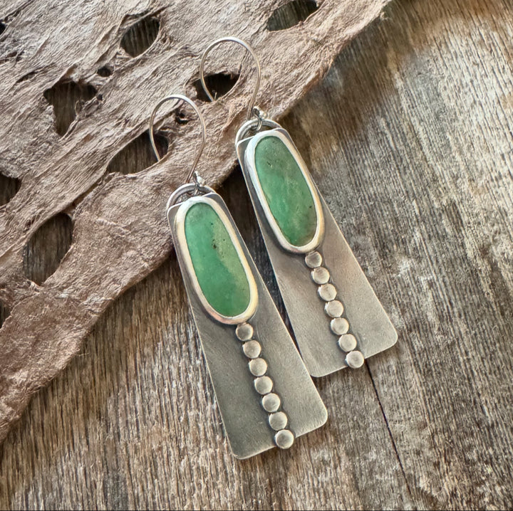 Close-up of handmade long turquoise earrings featuring green Elisa turquoise stones, sterling silver disc accents, and French hook earwires. Rustic wood and feather background .