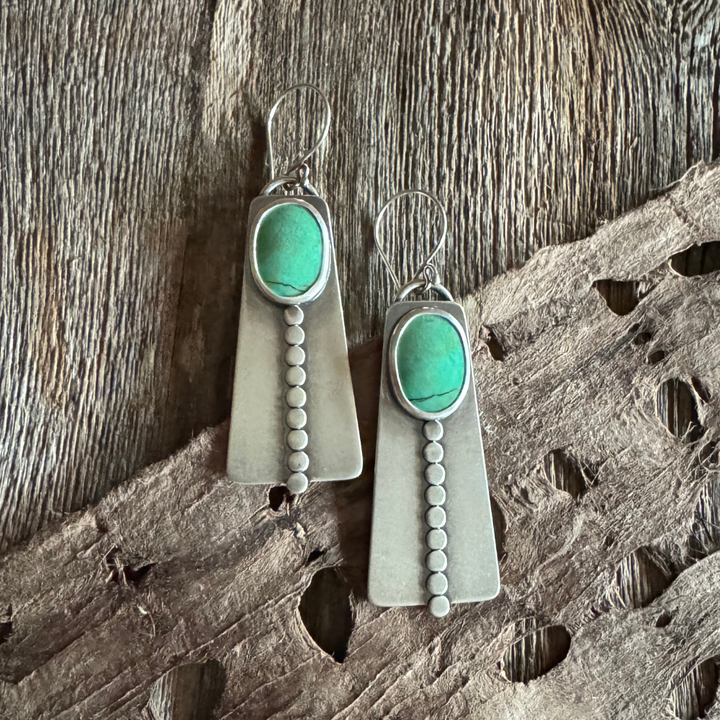 Close-up of handmade long turquoise earrings featuring green Elisa turquoise stones, sterling silver disc accents, and French hook earwires. Rustic wood and feather background .