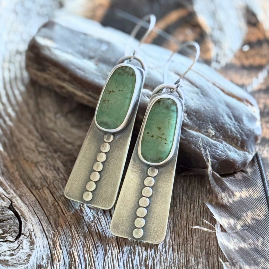Close-up of handmade long turquoise earrings featuring green Elisa turquoise stones, sterling silver disc accents, and French hook earwires. Rustic wood and feather background .