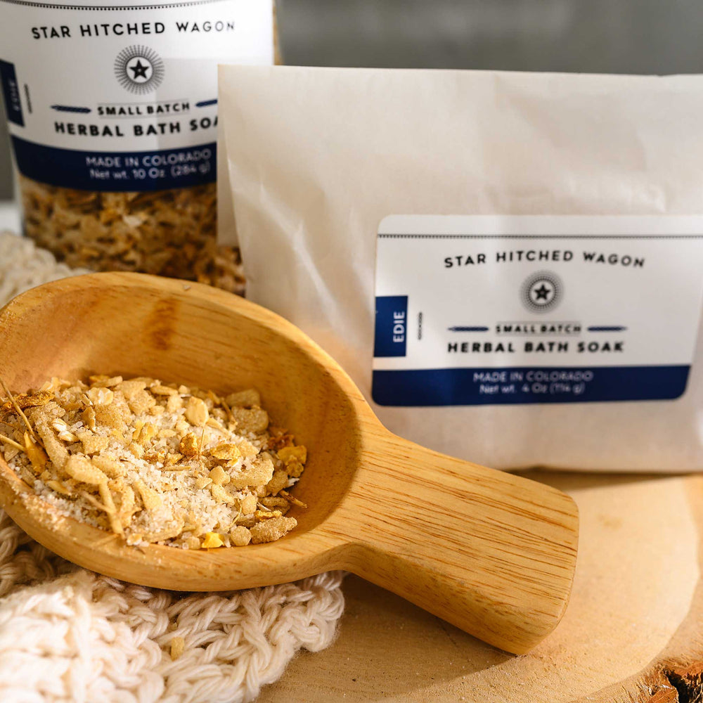 Wooden scoop filled with Edie Herbal Bath Soak next to full pouch and glass bottle on wooden slab next to a cream colored knit washcloth.