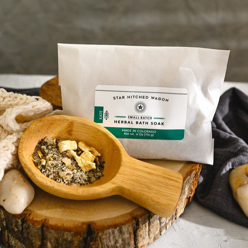 Wooden scoop filled with Kate Herbal Bath Soak next to full pouch on wooden slab next to a cream colored knit washcloth, a grey towel and pebbles.
