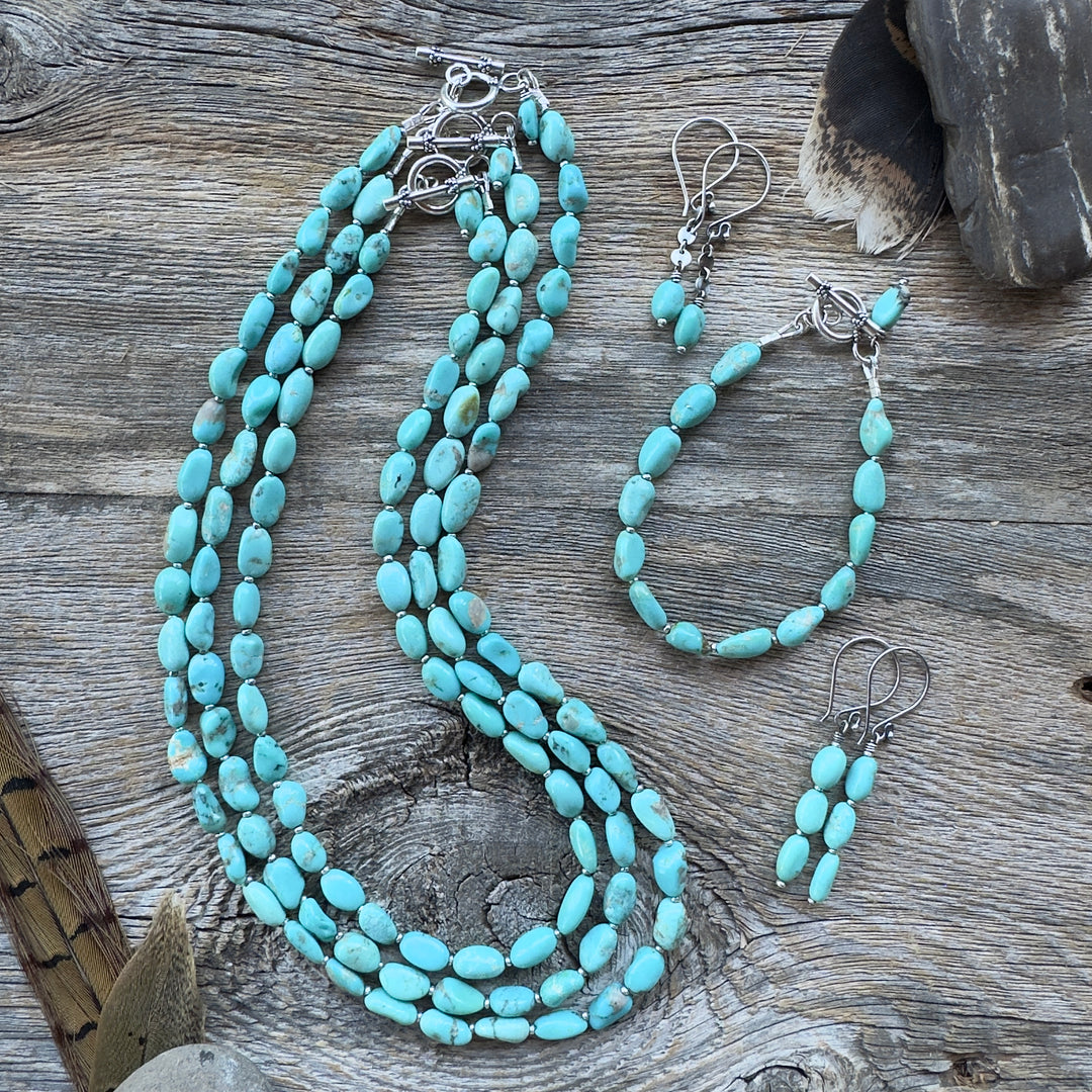 Graduated length turquoise nugget bead necklaces and bracelet with earrings, displayed on a rustic weathered wood surface with decorative feathers and stones in background.
