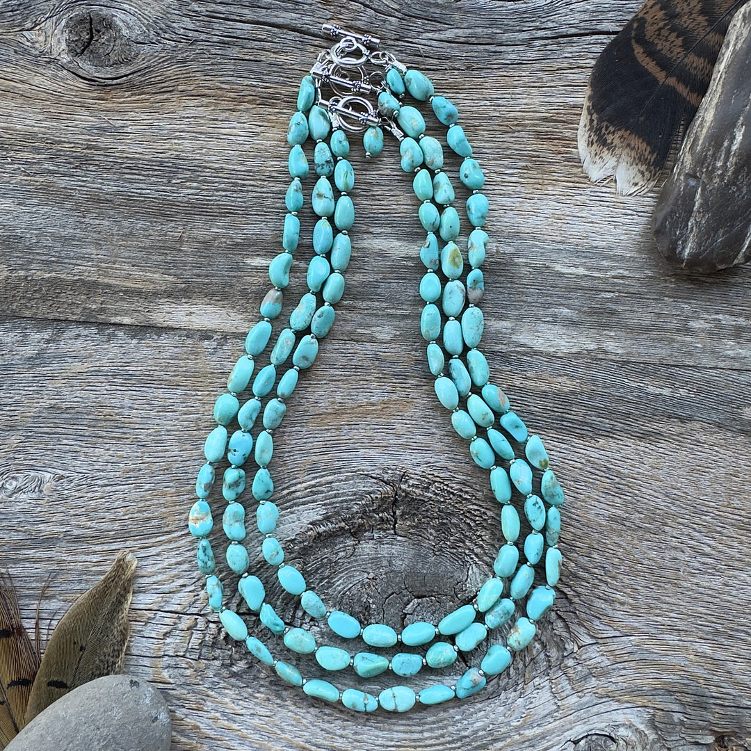 Three graduated length turquoise nugget bead necklaces with sterling silver clasps, displayed on a rustic weathered wood surface with decorative feathers and stones in background.