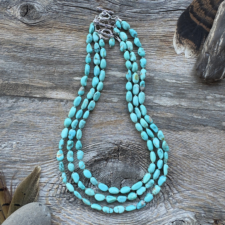 Three graduated length turquoise nugget bead necklaces with sterling silver clasps, displayed on a rustic weathered wood surface with decorative feathers and stones in background.