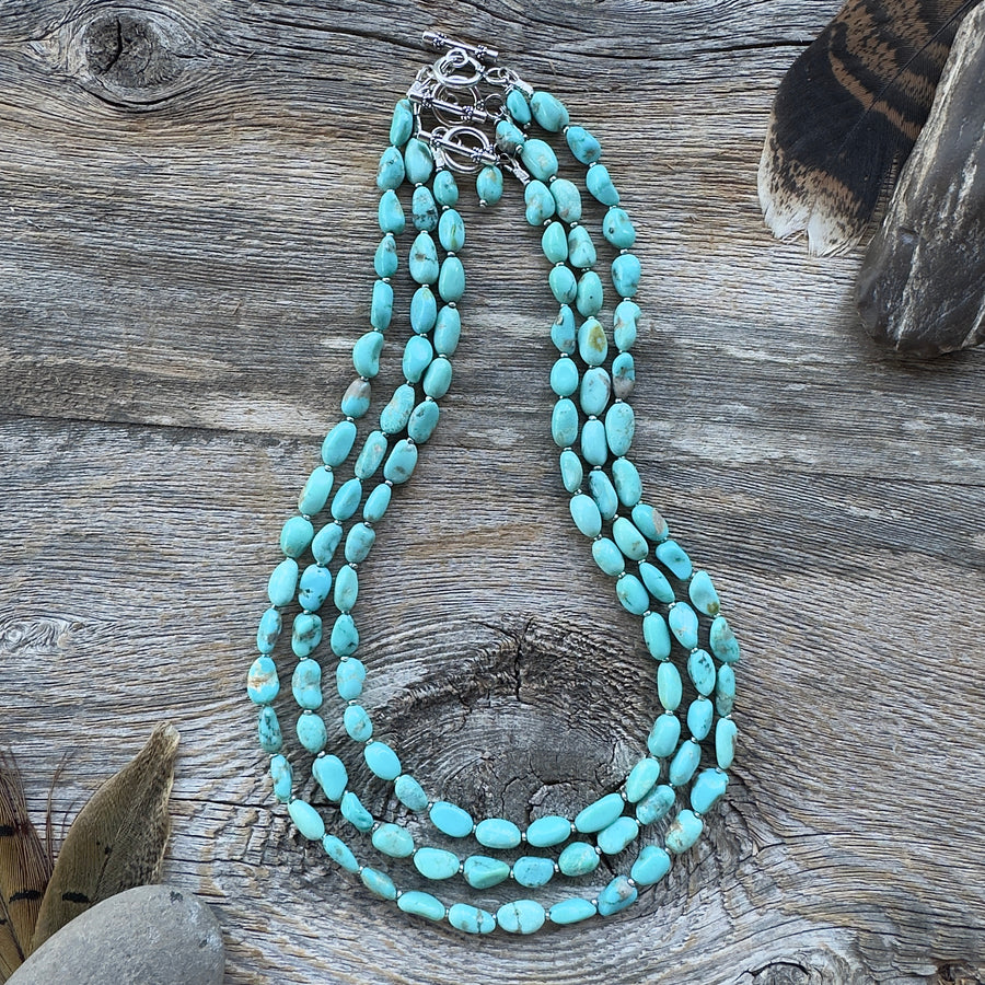 Three graduated length turquoise nugget bead necklaces with sterling silver clasps, displayed on a rustic weathered wood surface with decorative feathers and stones in background.