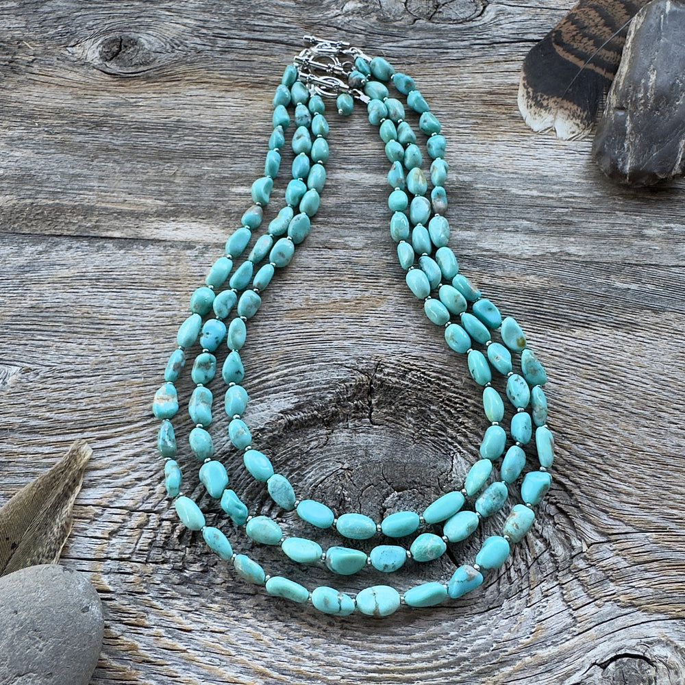 Three graduated length turquoise nugget bead necklaces with sterling silver clasps, displayed on a rustic weathered wood surface with decorative feathers and stones in background.