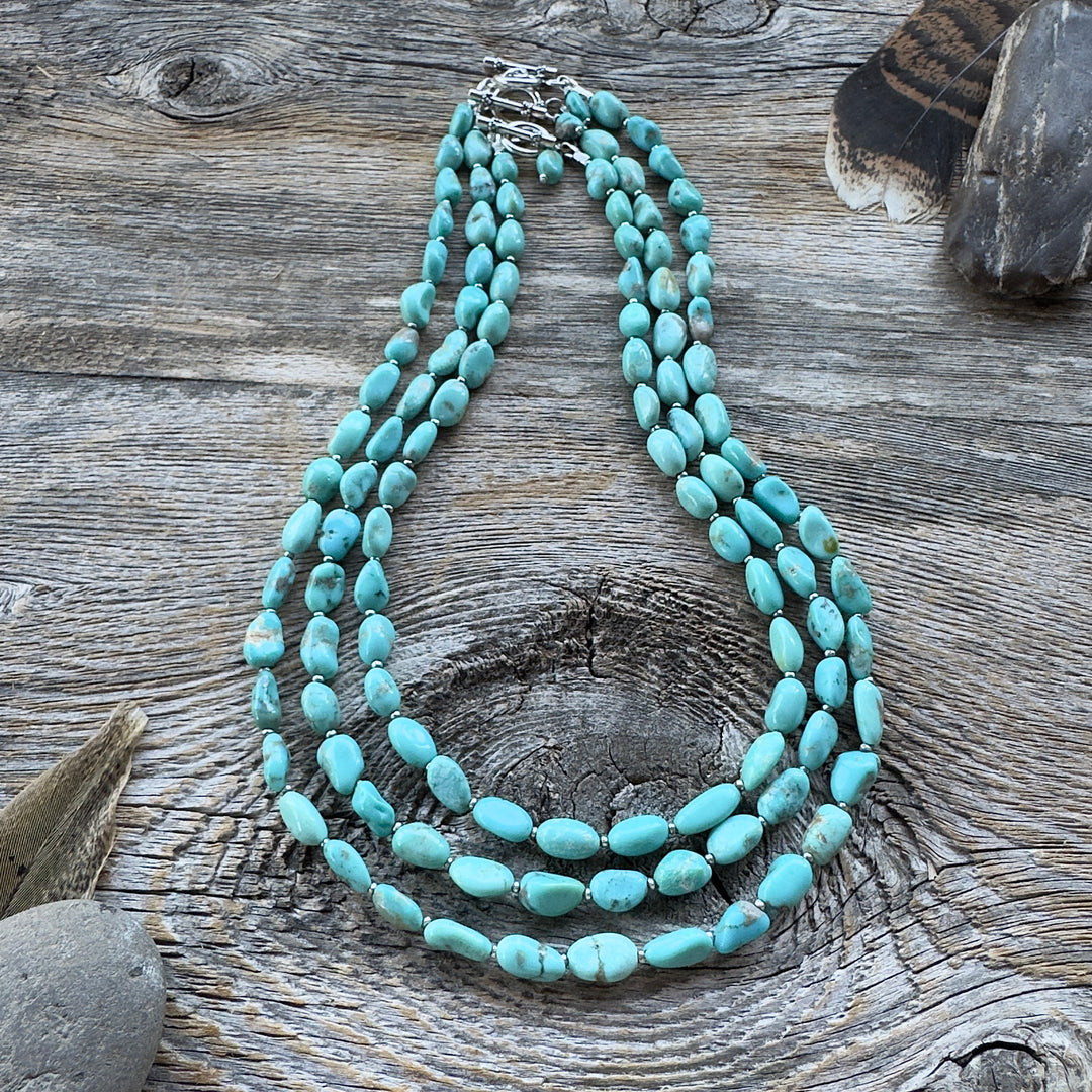 Three graduated length turquoise nugget bead necklaces with sterling silver clasps, displayed on a rustic weathered wood surface with decorative feathers and stones in background.