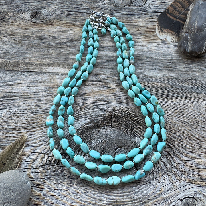 Three graduated length turquoise nugget bead necklaces with sterling silver clasps, displayed on a rustic weathered wood surface with decorative feathers and stones in background.
