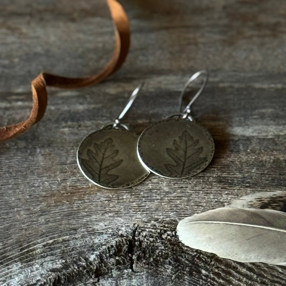 Silver leaf earrings on a wooden surface with a feather and leather lace.