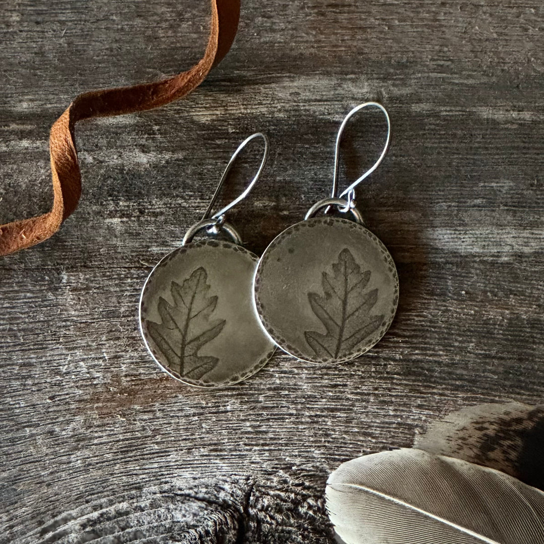 Silver leaf-patterned earrings on a wooden surface with a feather and leather lace.