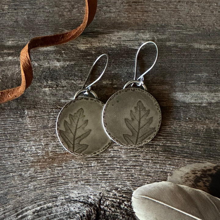 Silver leaf-patterned earrings on a wooden surface with a feather and leather lace.