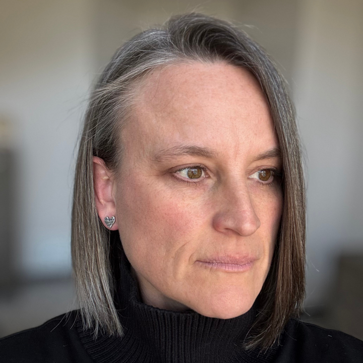 Woman with short hair wearing a black turtleneck sweater and sterling silver script heart post earrings against a blurred background.