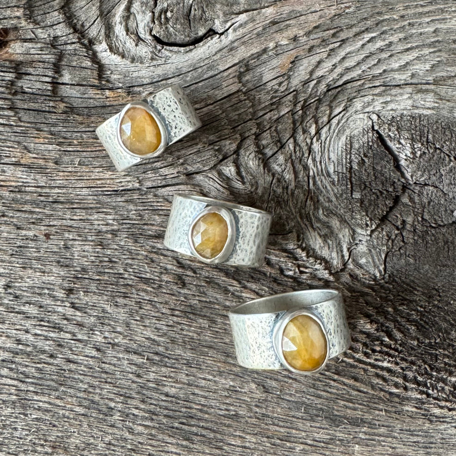 Sterling silver ring with yellow sapphire featuring a faceted golden stone on a wide hammered band with brushed patina finish on rustic wood backdrop.