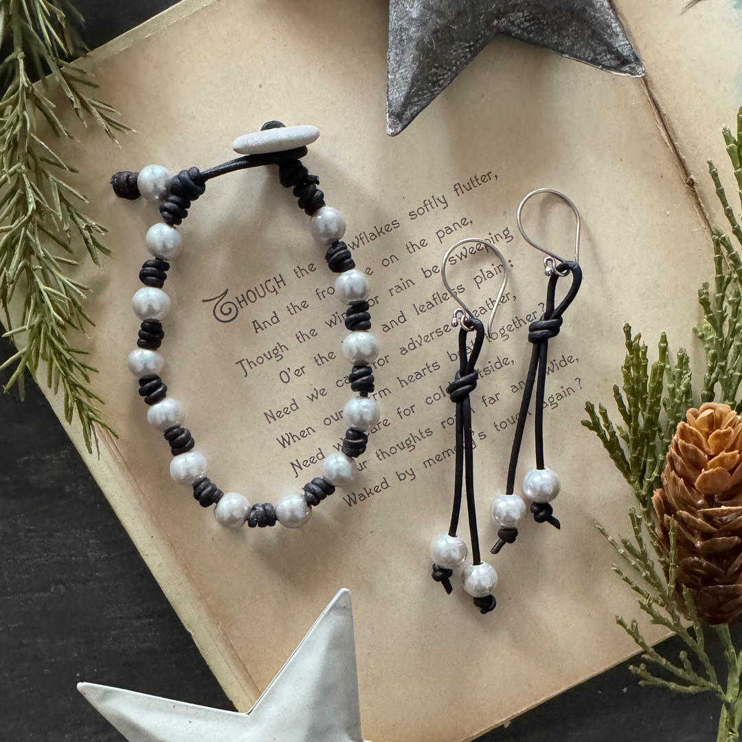 Black leather and white pearl bracelet with pebble button closure shown beside matching long leather and pearl dangle earrings on a rustic holiday background.