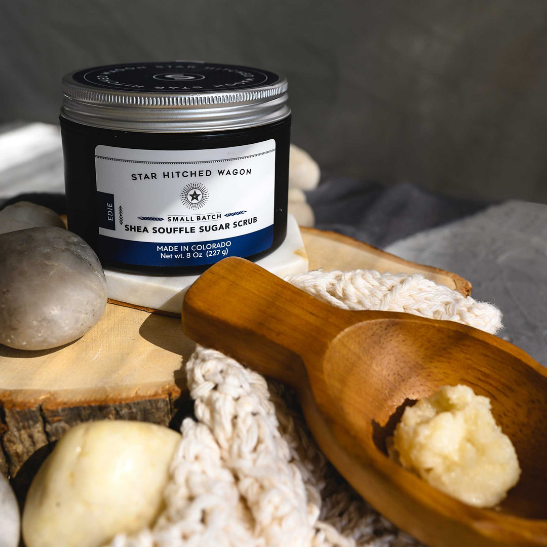 An 8 ounce jar of Edie Shea Souffle Sugar Scrub is displayed on a wooden surface, accompanied by a pebbles, wooden scoop and a knit wash cloth.