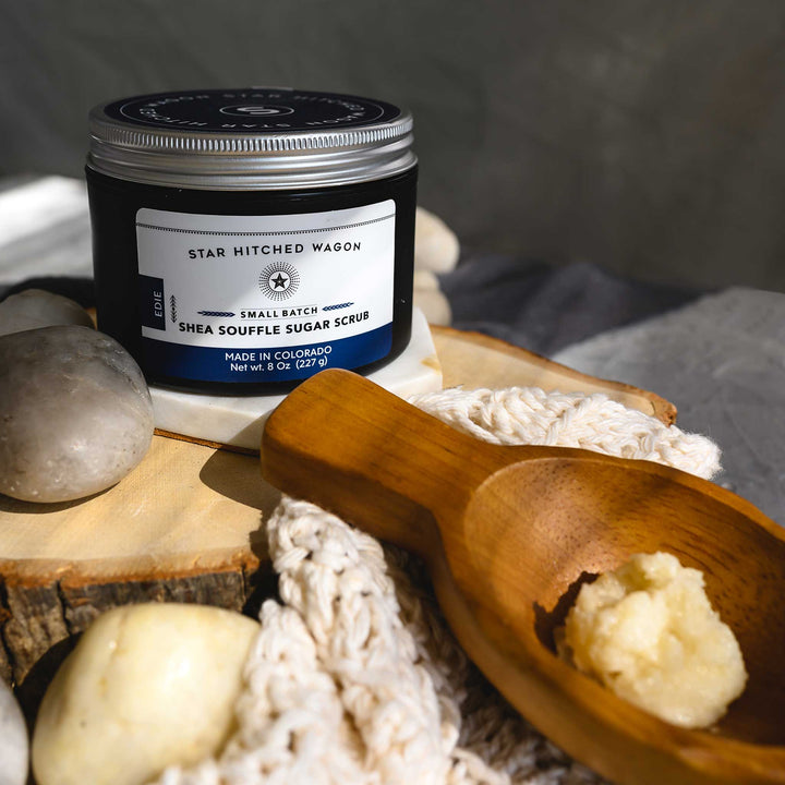 An 8 ounce jar of Edie Shea Souffle Sugar Scrub is displayed on a wooden surface, accompanied by a pebbles, wooden scoop and a knit wash cloth.