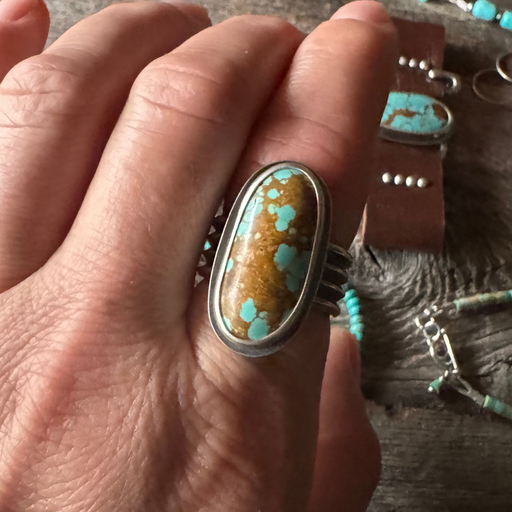 Hand wearing a turquoise stone ring with jewelry in the background.