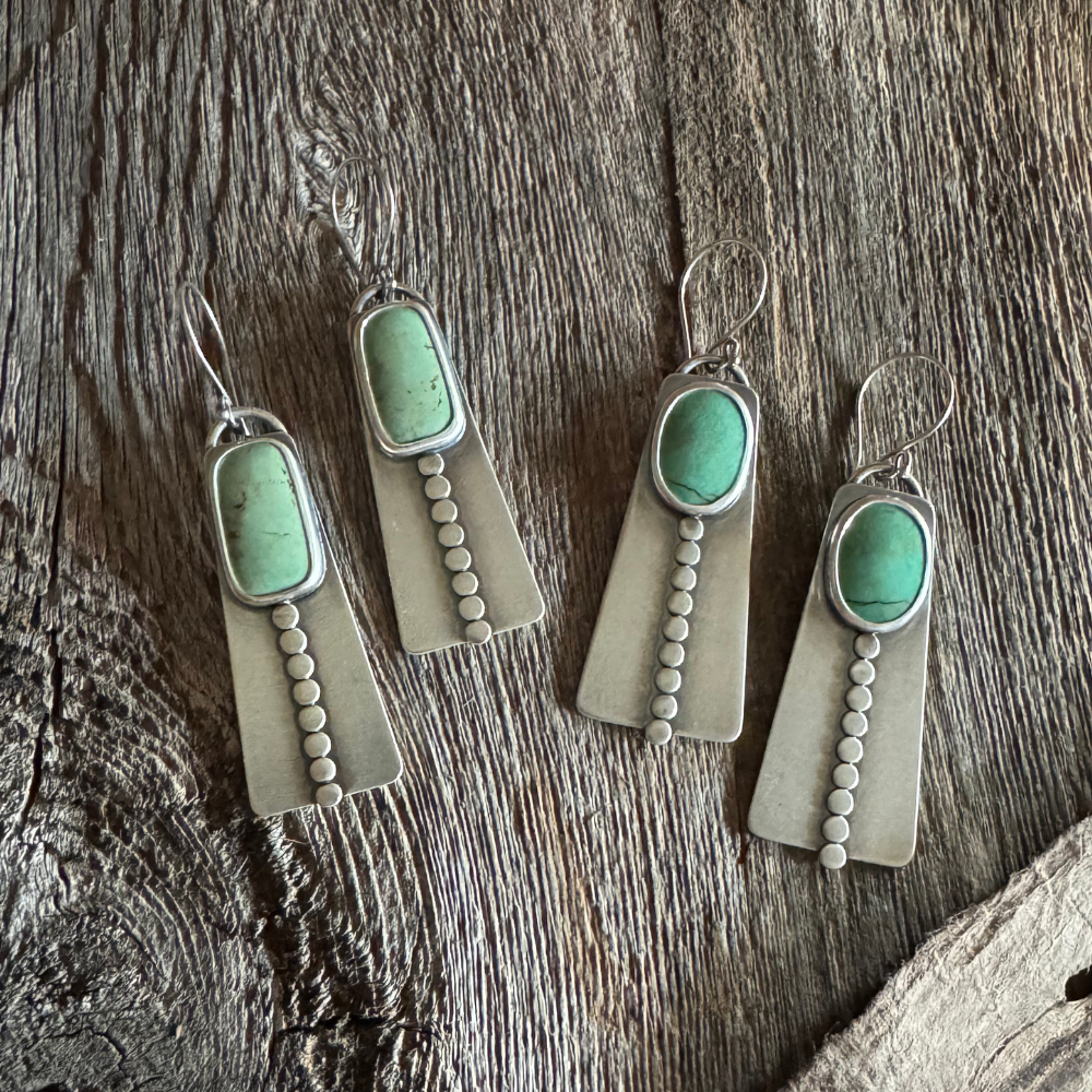 Close-up of two pairs of handmade long turquoise earrings featuring green Elisa turquoise stones, sterling silver disc accents, and French hook earwires. Rustic wood background l.