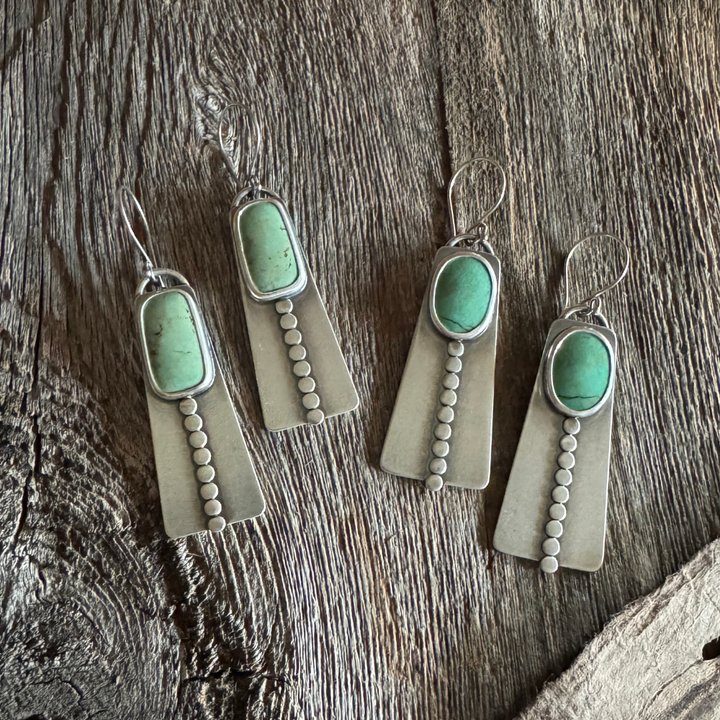 Close-up of two pairs of handmade long turquoise earrings featuring green Elisa turquoise stones, sterling silver disc accents, and French hook earwires. Rustic wood background l.