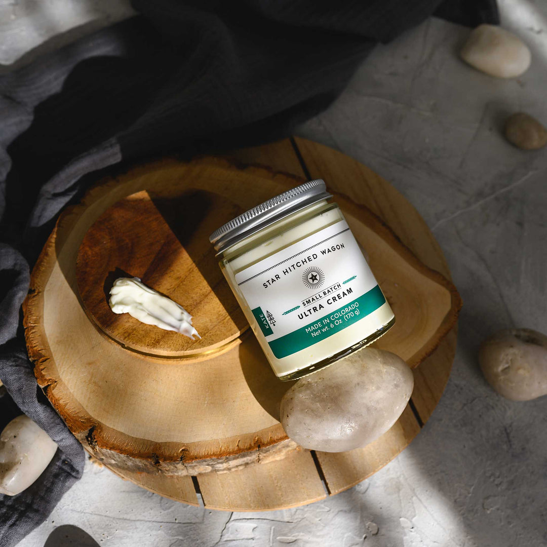 Kate Ultra Cream in a glass jar with an aluminum screw top lid, displayed on a wooden surface with a grey cloth and pebbles in the background.