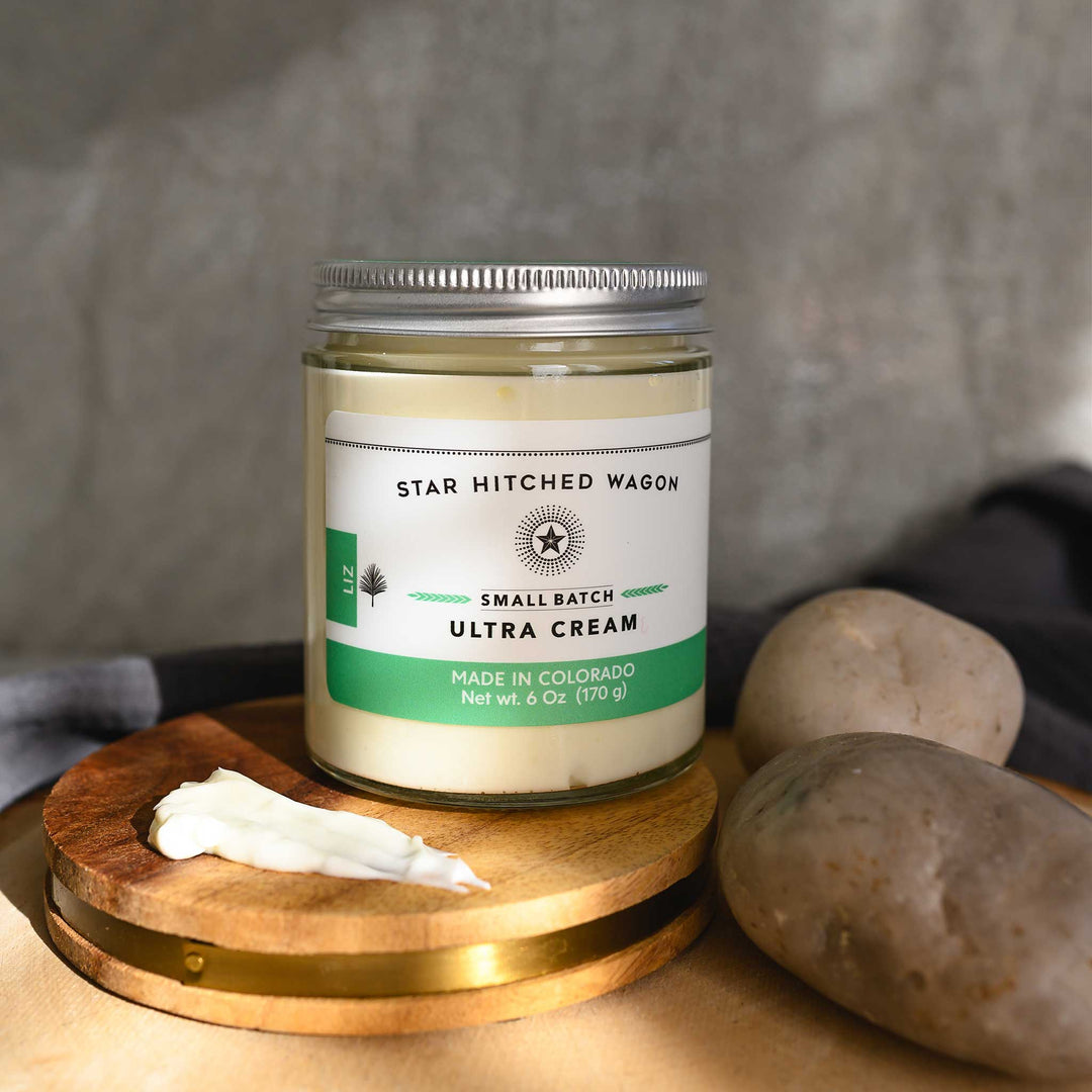 Liz Ultra Cream in a glass jar with an aluminum screw top lid, displayed on a wooden surface with a grey cloth and pebbles in the background.