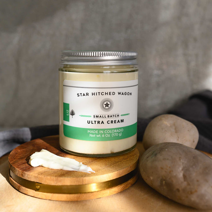 Liz Ultra Cream in a glass jar with an aluminum screw top lid, displayed on a wooden surface with a grey cloth and pebbles in the background.