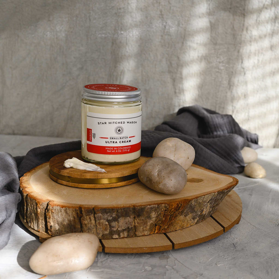 Margo Ultra Cream in a glass jar with an aluminum screw top lid, displayed on a wooden surface with a grey cloth and pebbles in the background.