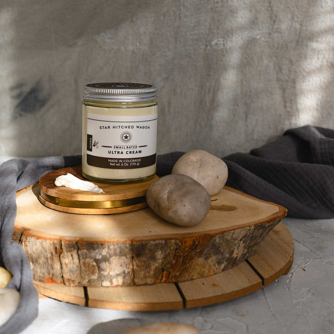 Nora Ultra Cream in a glass jar with an aluminum screw top lid, displayed on a wooden surface with a grey cloth and pebbles in the background.