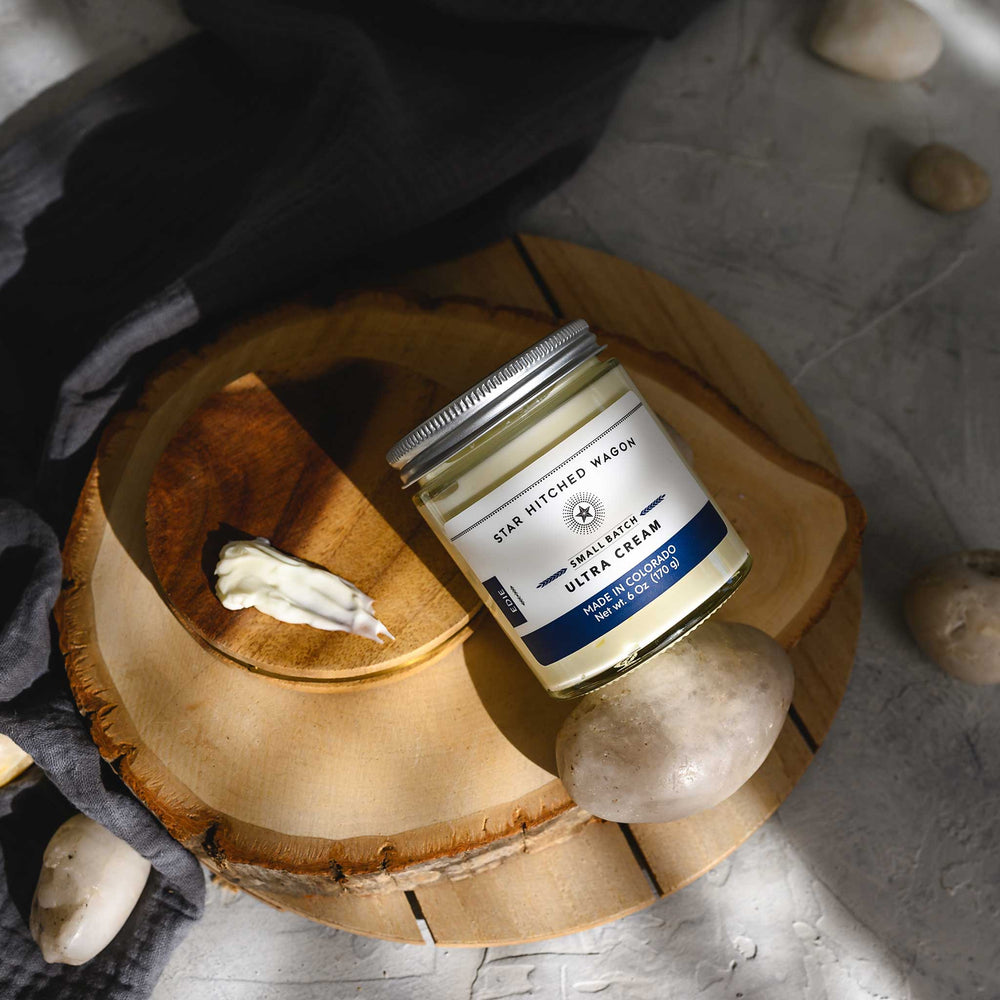 Edie Ultra Cream in a glass jar with an aluminum screw top lid, displayed on a wooden surface with a grey cloth and pebbles in the background.