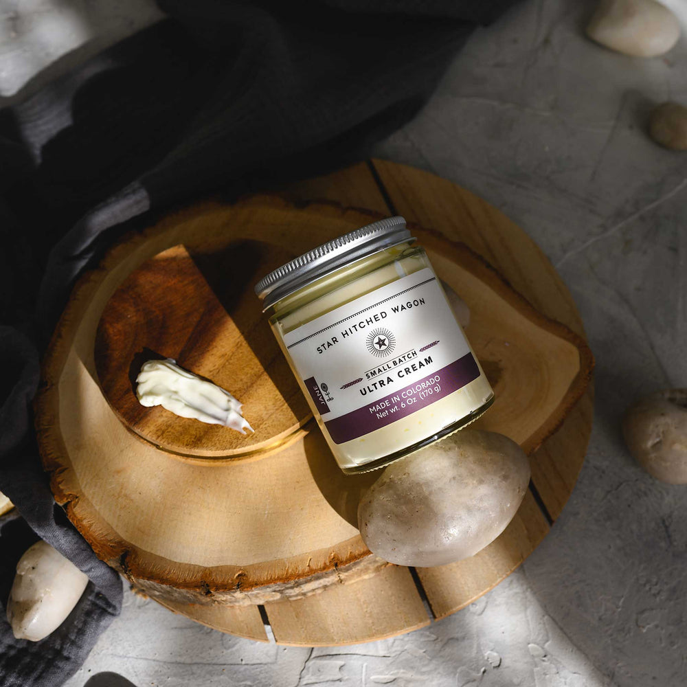 Jane Ultra Cream in a glass jar placed on a wooden disc, alongside pebbles and a grey towel. 