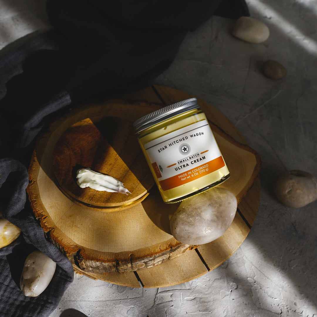 Jess Ultra Cream in a glass jar placed on a wooden platform, pebbles and grey towel in foreground and background.