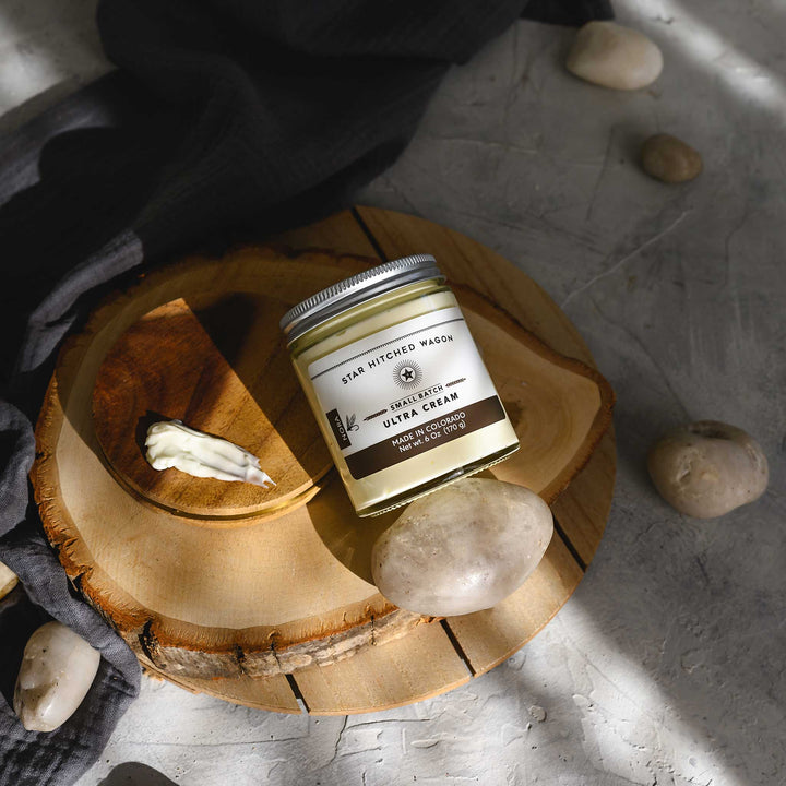 Nora Ultra Cream in a glass jar with an aluminum screw top lid, displayed on a wooden surface with a grey cloth and pebbles in the background.
