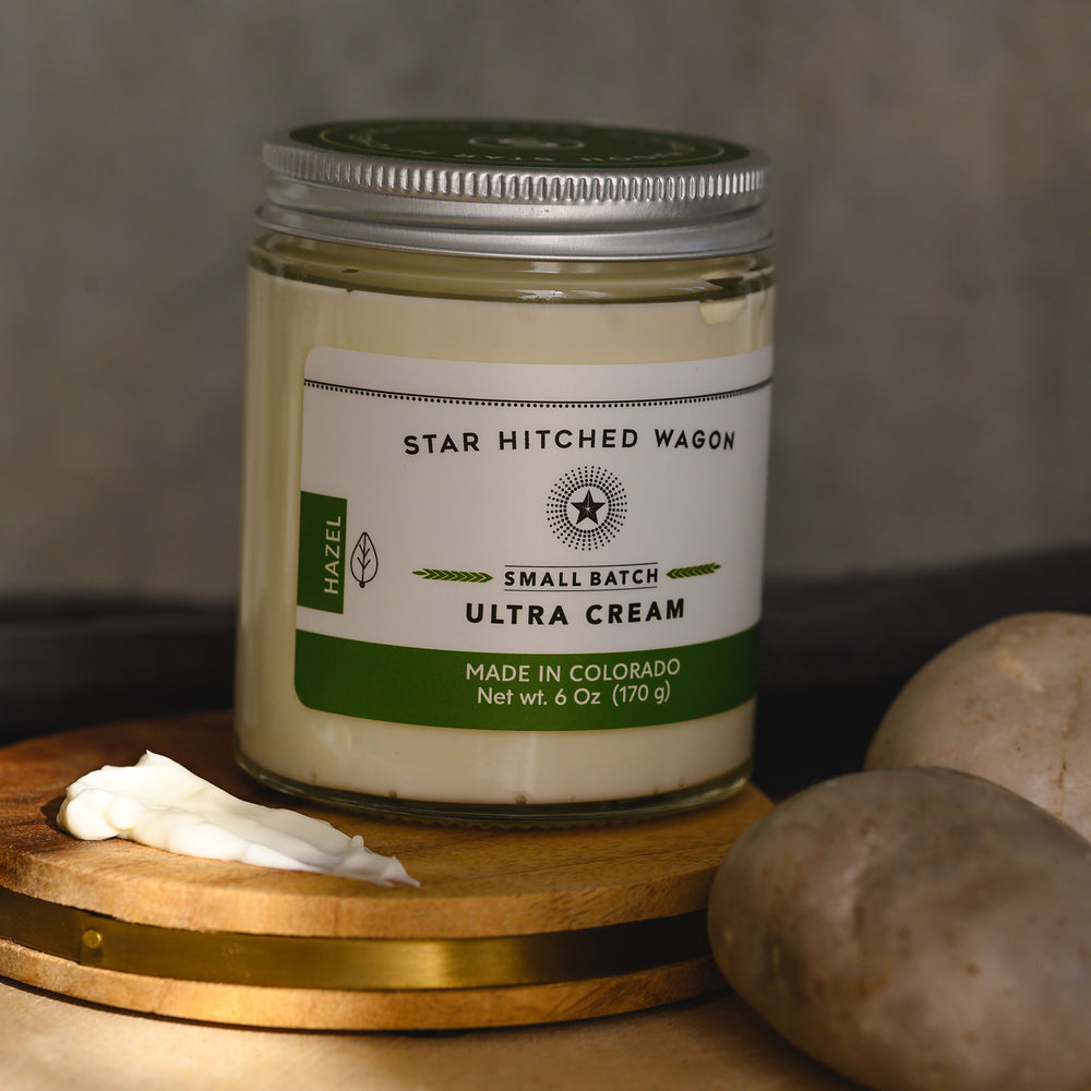 Hazel Ultra Cream in a glass jar with an aluminum screw top lid, placed on a wooden surface with pebbles and grey towel in the background.