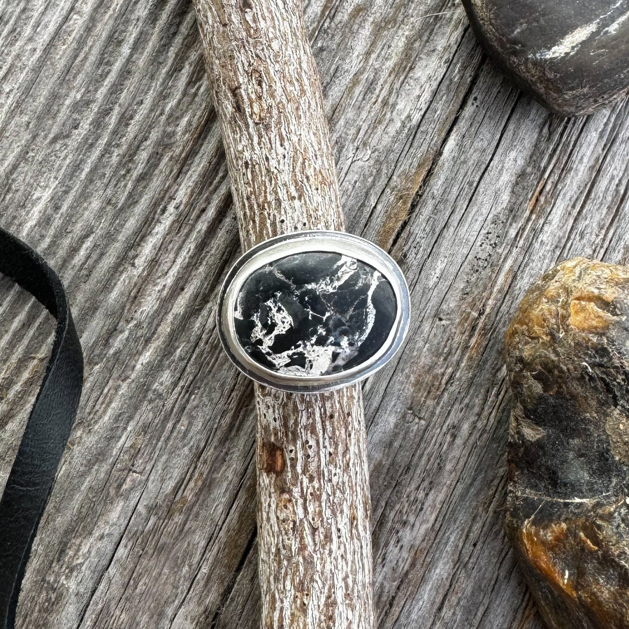 A white buffalo turquoise ring design on a wooden surface, accompanied by a black leather strap, twigs and rocks.
