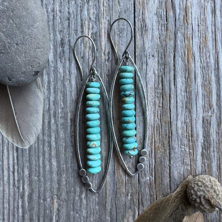 A pair of long sterling silver and Kingman turquoise earrings with open leaf and stacked turquoise rondelle design , displayed on a rustic weathered wood background with decorative feathers and a stone.