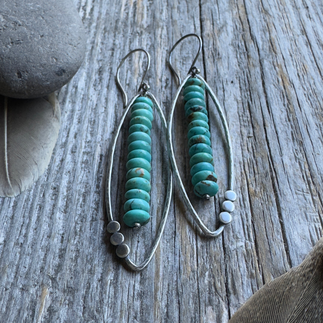 A pair of long sterling silver and Kingman turquoise earrings with open leaf and stacked turquoise rondelle design , displayed on a rustic weathered wood background with decorative feathers and a stone.
