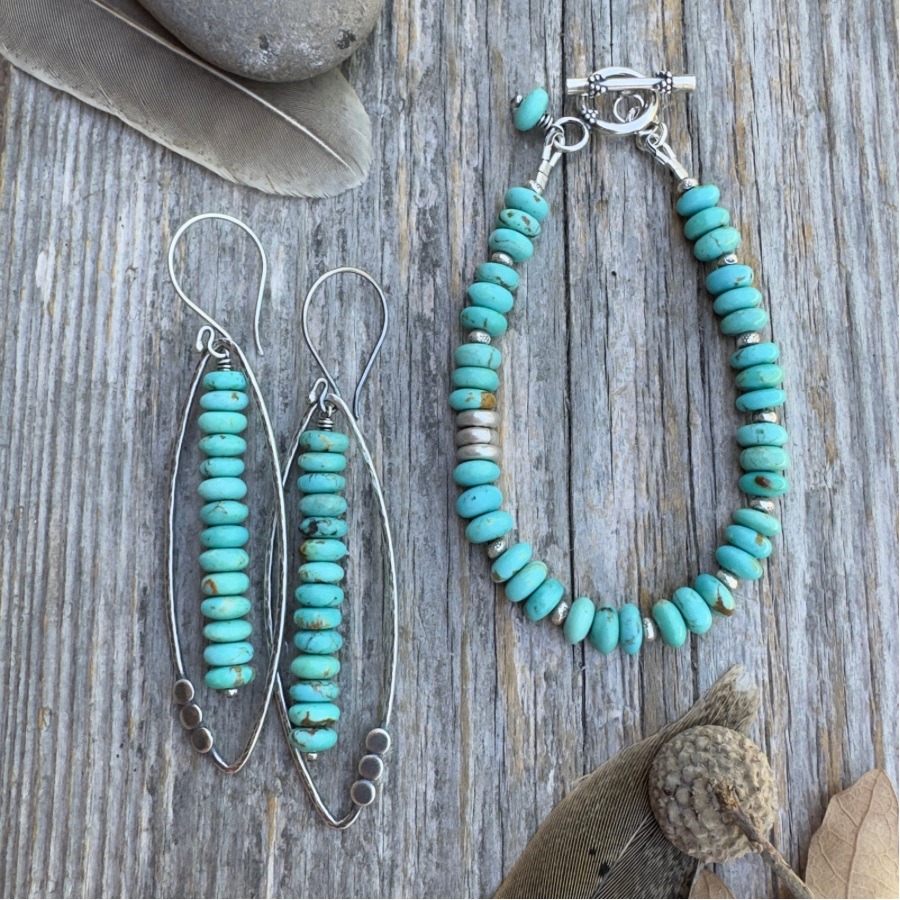 A pair of long sterling silver and Kingman turquoise earrings with open leaf and stacked turquoise rondelle design alongside a bracelet with the same stacked turquoise rondelles with silver, both displayed on a rustic weathered wood background with decorative feathers and a stone.