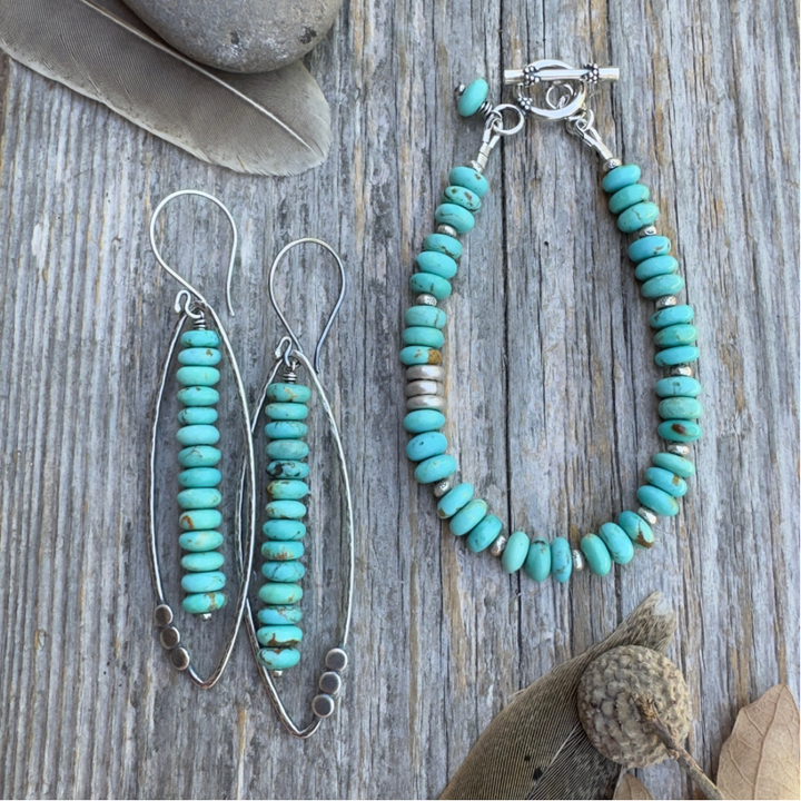 A pair of long sterling silver and Kingman turquoise earrings with open leaf and stacked turquoise rondelle design alongside a bracelet with the same stacked turquoise rondelles with silver, both displayed on a rustic weathered wood background with decorative feathers and a stone.