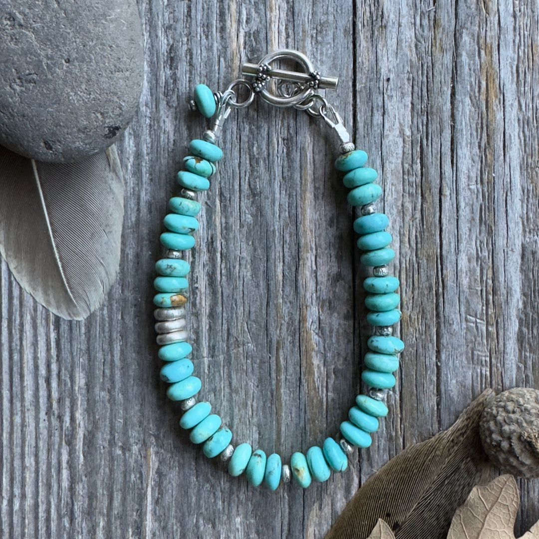 A beaded bracelet with blue Kingman turquoise rondelle beads and small Thai silver spacer beads, arranged in an oval shape on a weathered gray wood surface with decorative stones and dried leaves nearby.
