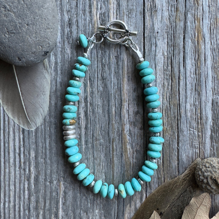 A beaded bracelet with blue Kingman turquoise rondelle beads and small Thai silver spacer beads, arranged in an oval shape on a weathered gray wood surface with decorative stones and dried leaves nearby.